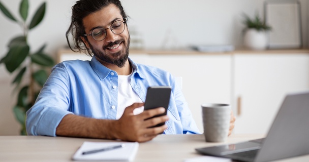 Happy Indian man using his phone and laptop drinking coffee