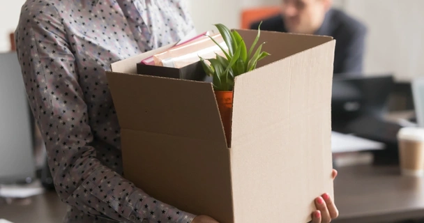 Woman in spotty shirt carrying a cardboard box of belongings from an office. A man works in the background