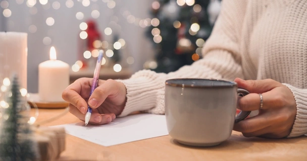 Woman in white sweater is writing a list while drinking from a mug. There's a Christmas tree, candles and twinkly fairy lights in the background.