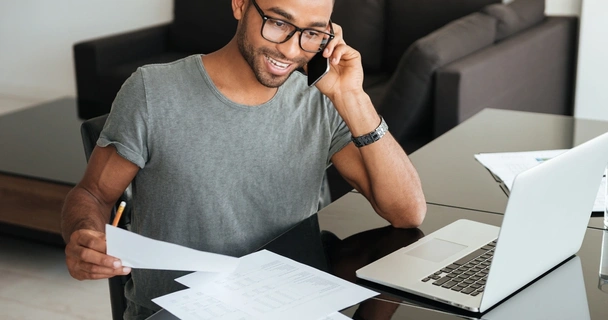 smiling man on the phone sat at a desk with his laptop and paperwork