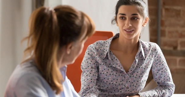 A dark haired lady in a spotted lilac shirt talks to a red haired lady with her back to the camera. They're both sitting at a desk.