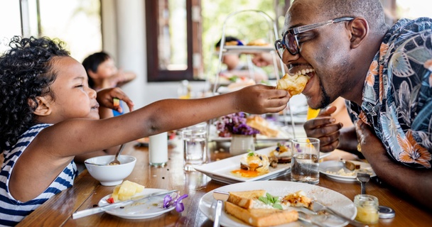 A daughter is feeding her father sandwiches in a cafe