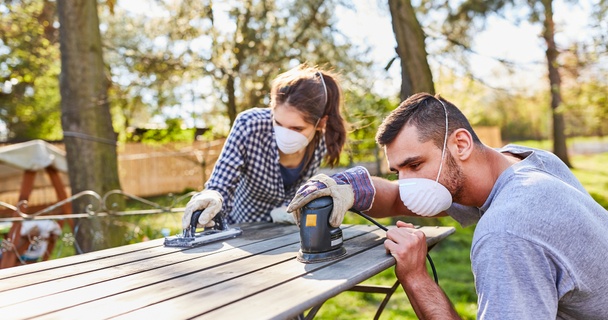 Couple wearing dust masks sand down a wooden table outdoors