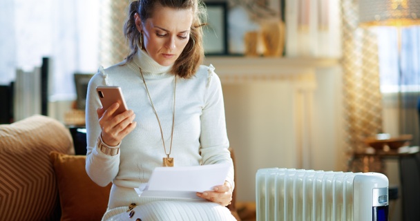 Woman wrapped up next to a radiator paying her energy bills on her mobile.