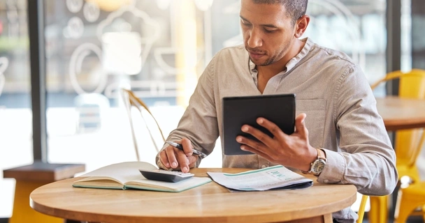 man sat at a table in a cafe looking at his laptop and calculator