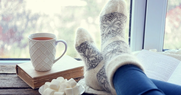 woman with feet up on window sill with a hot drink, book and marshmallows