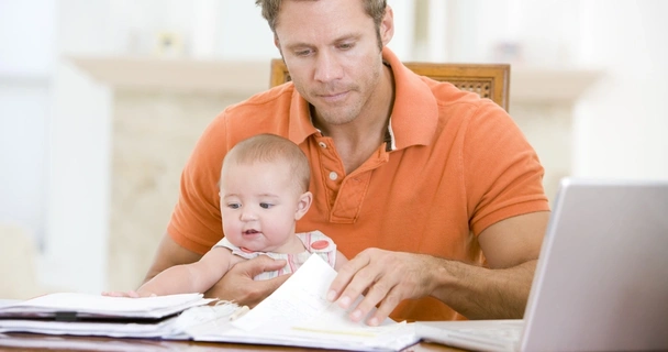 Father sat with baby at a table looking at paperwork with the laptop open