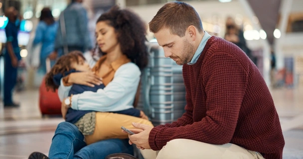 Family of three sat on the floor at the airport with the father looking at his phone.