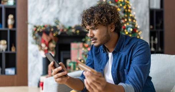 Man sat in living room which is decorated for Christmas. He's holding his phone and credit card and looking concerned.