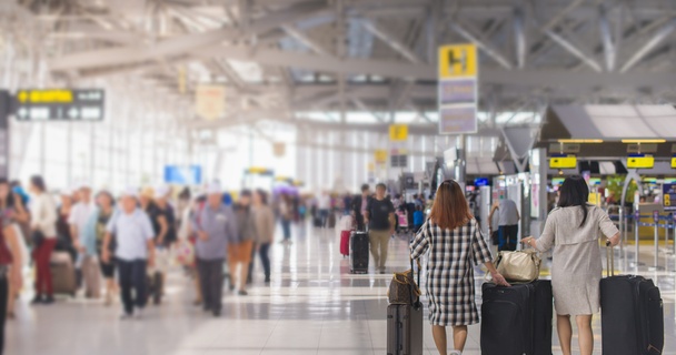 two women, one in a checked dress and one in a cream dress, wheel their suitcases through a busy airport terminal