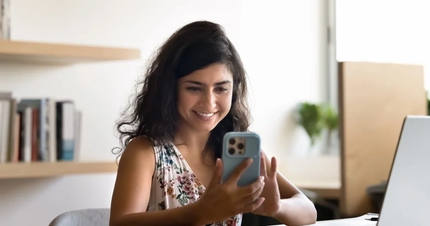 A woman sat at her desk is checking her credit score on her mobile phone and smiling