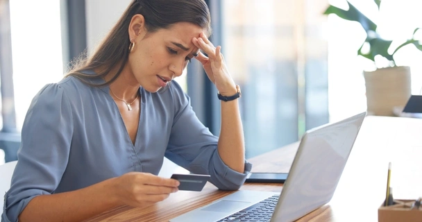 Woman sat at a laptop and looking at her credit card with a concerned expression