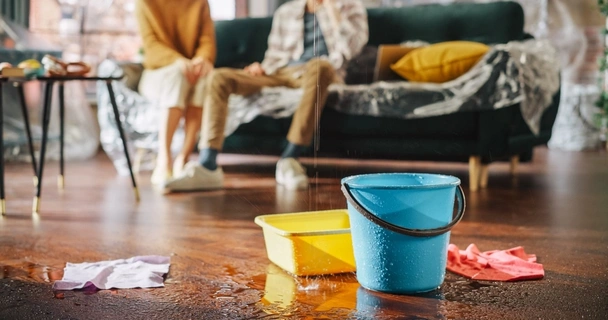 Couple sat on the sofa, with a close up of buckets on the living room floor catching water from a roof leak