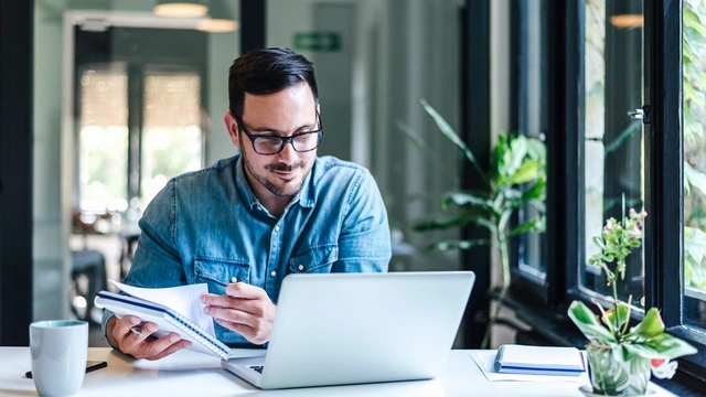 man looking at his computer while holding a notebook