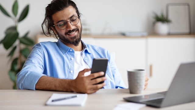 Happy Indian man using his phone and laptop drinking coffee