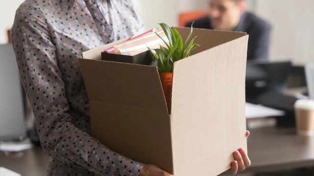 Woman in spotty shirt carrying a cardboard box of belongings from an office. A man works in the background