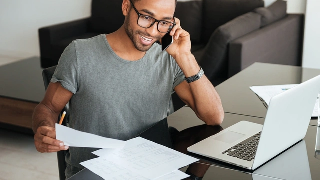 smiling man on the phone sat at a desk with his laptop and paperwork