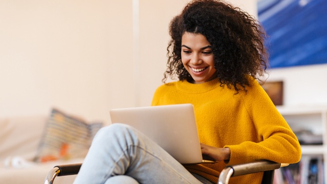 Lady with curly hair in yellow jumper smiling while using a laptop