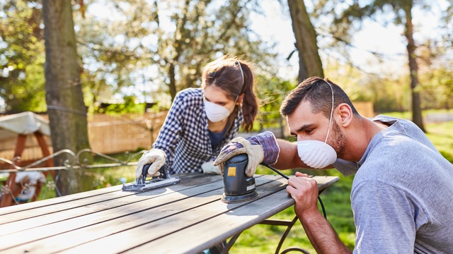 Couple wearing dust masks sand down a wooden table outdoors