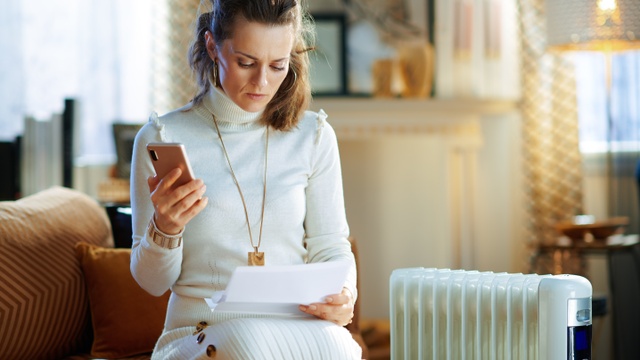 Woman wrapped up next to a radiator paying her energy bills on her mobile.