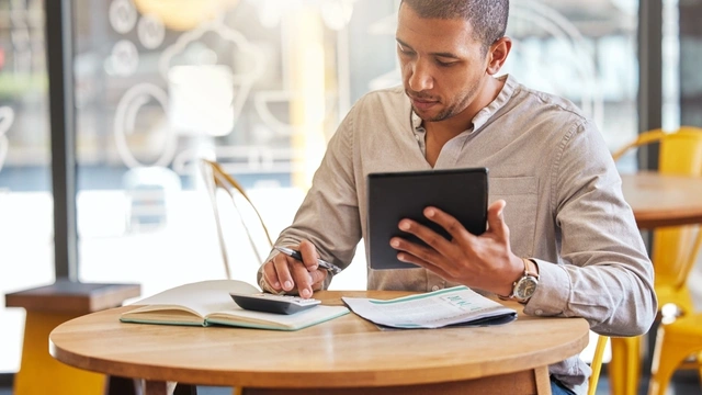 man sat at a table in a cafe looking at his laptop and calculator