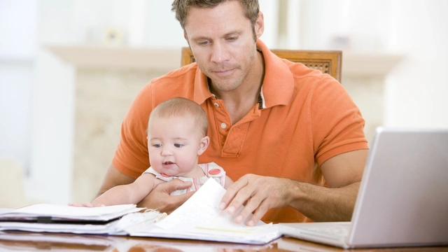 Father sat with baby at a table looking at paperwork with the laptop open