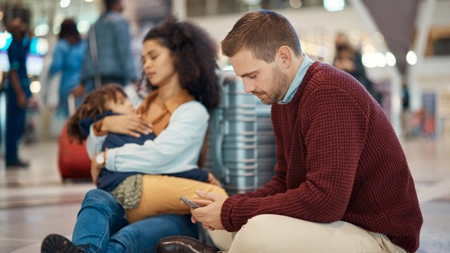 Family of three sat on the floor at the airport with the father looking at his phone.