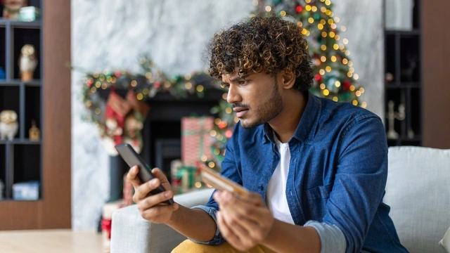 Man sat in living room which is decorated for Christmas. He's holding his phone and credit card and looking concerned.