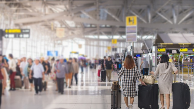 two women, one in a checked dress and one in a cream dress, wheel their suitcases through a busy airport terminal