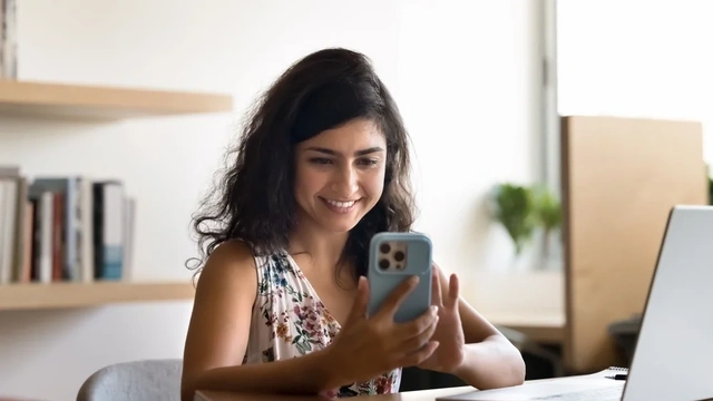 A woman sat at her desk is checking her credit score on her mobile phone and smiling