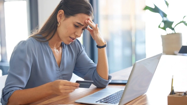 Woman sat at a laptop and looking at her credit card with a concerned expression