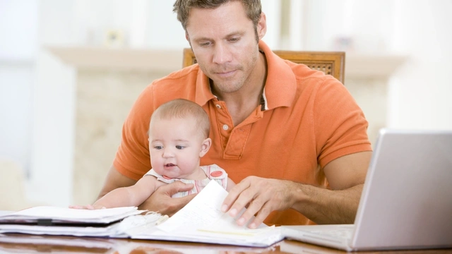 Father sat with baby at a table looking at paperwork with the laptop open