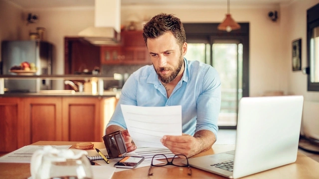 Man sat at the table looking through documents with his wallet and laptop open