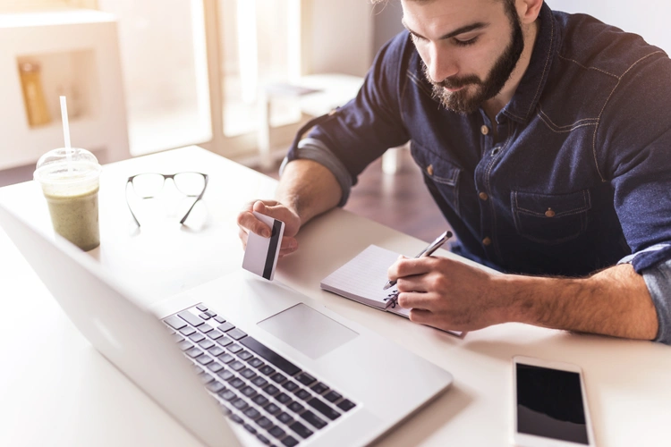 A man holding a credit card in front of a laptop while writing down a note