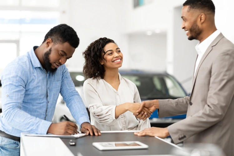 man signing car agreement and lady shaking hands with car salesman