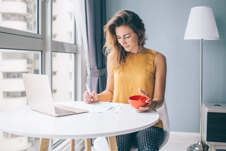 Woman writing in a notebook with a laptop in front of her