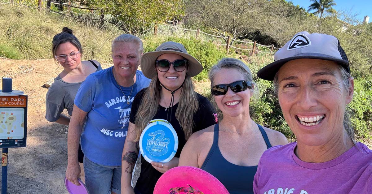 A group of women hold discs and smile near a disc golf tee