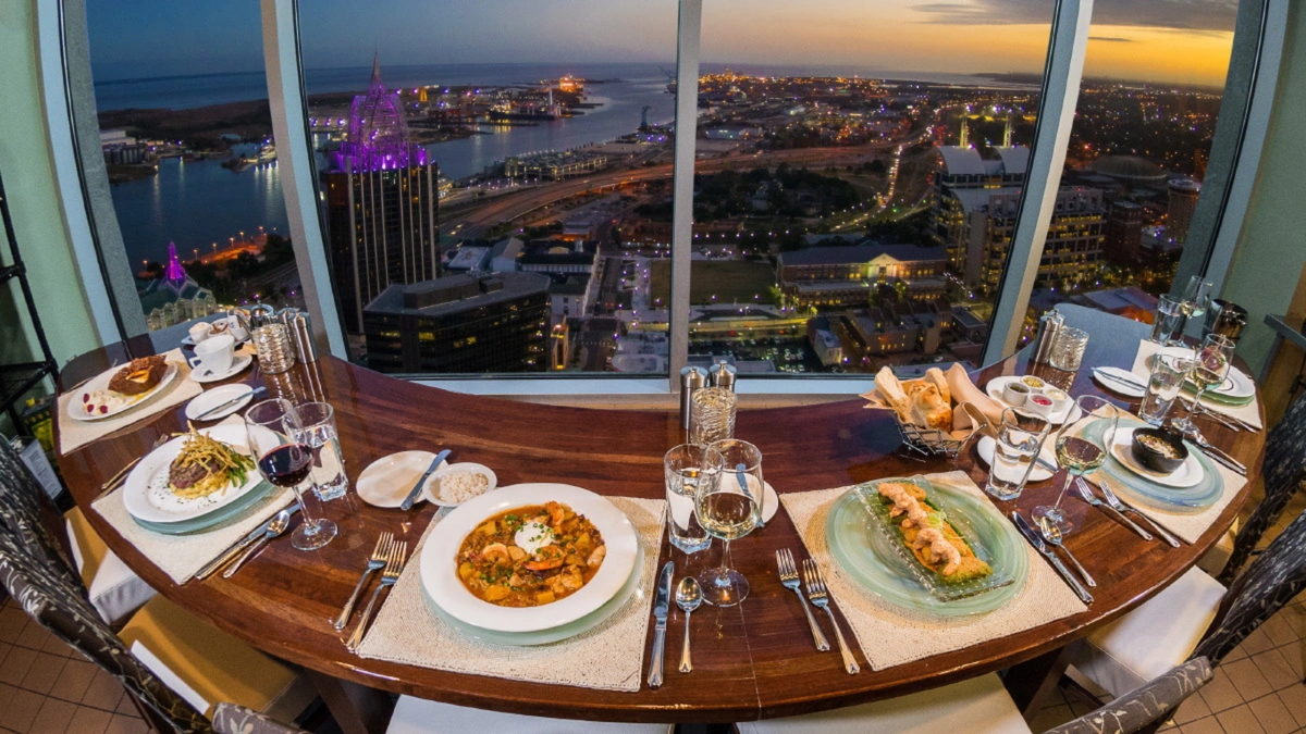 A curved dining table set for six with gourmet dishes, overlooking a city with tall buildings and a waterfront at sunset.