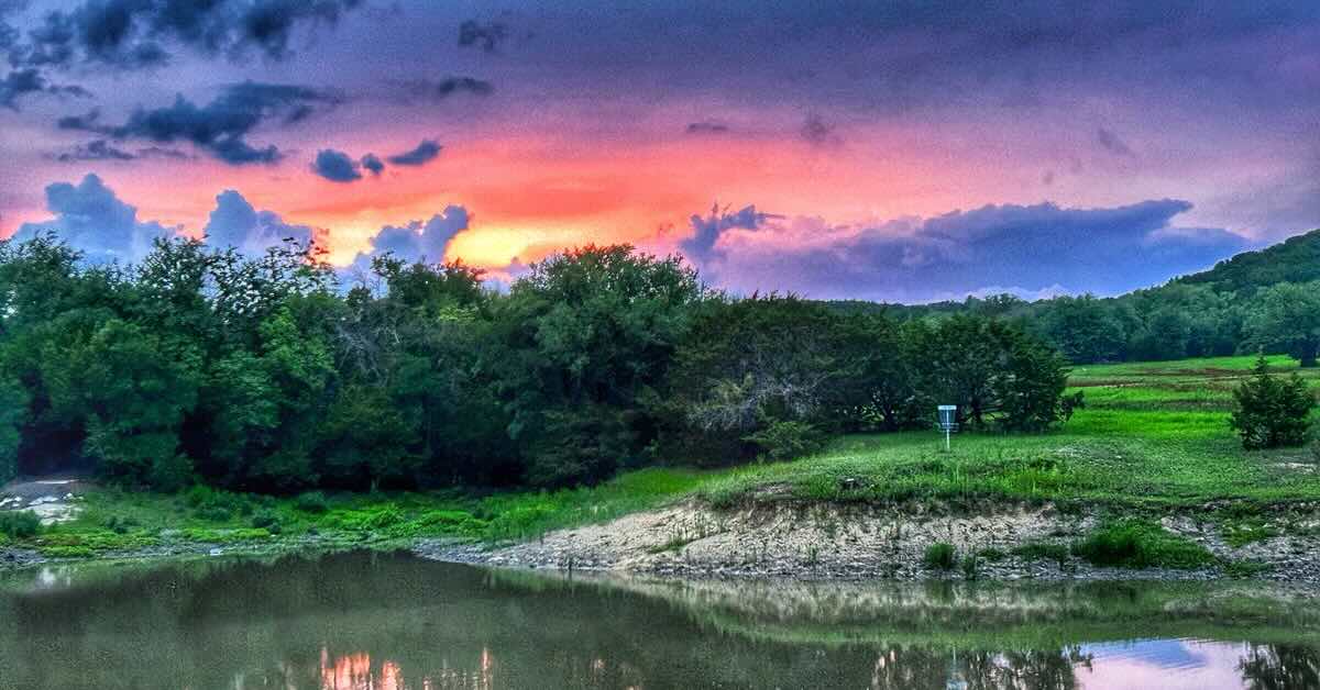 An area of mown grass and small trees and brush with gentle hills, sunset, and a disc golf basket