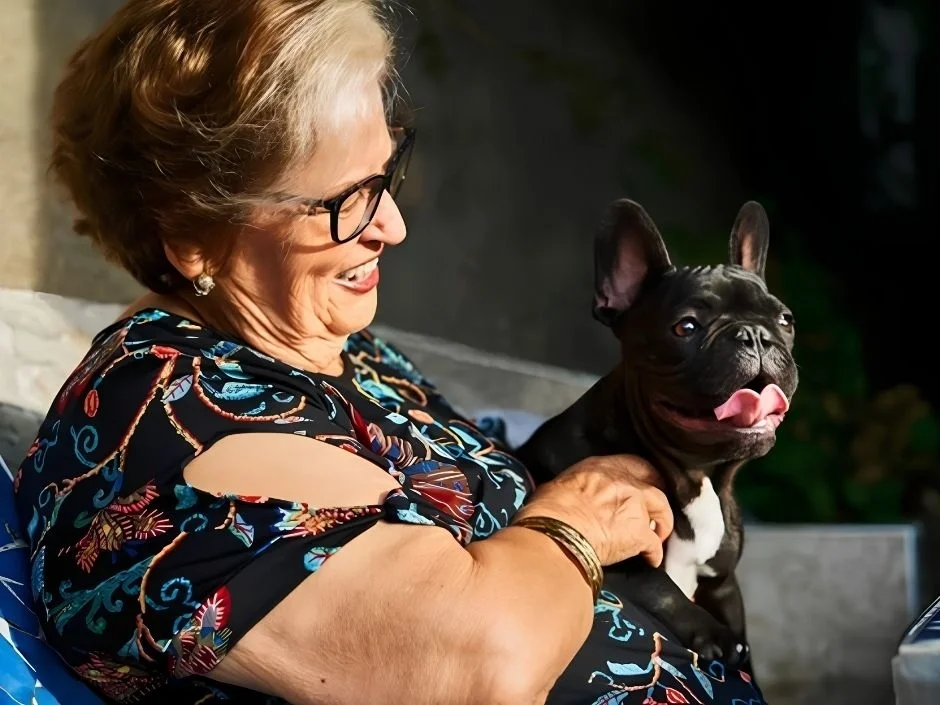 Retired woman in a floral dress sitting outdoors with a black French Bulldog puppy on her lap