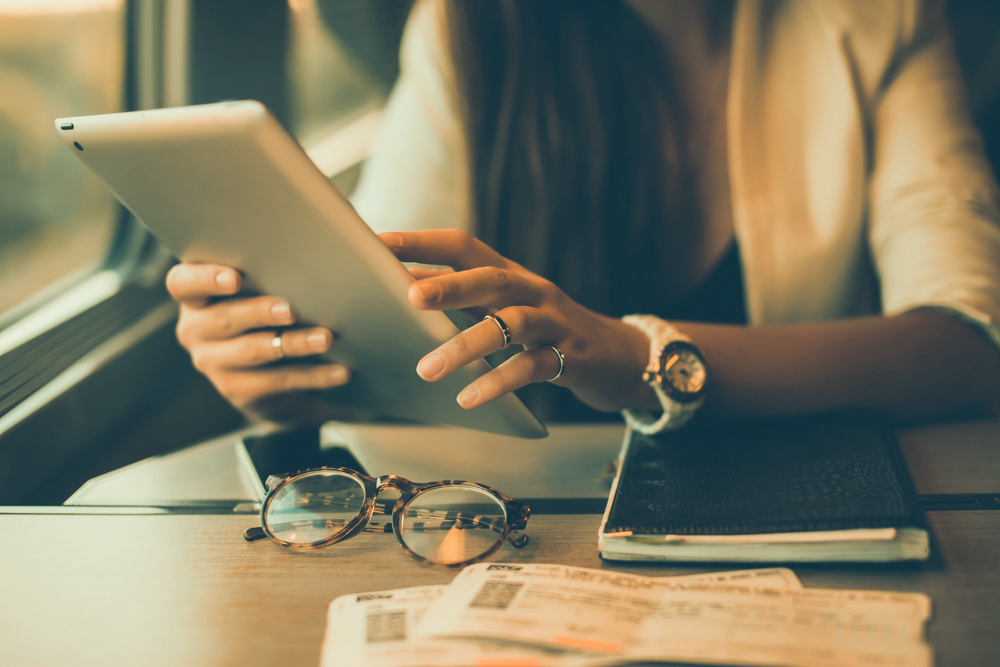 Photo of a person holding an iPad above a table that has tickets, a notebook, and glasses