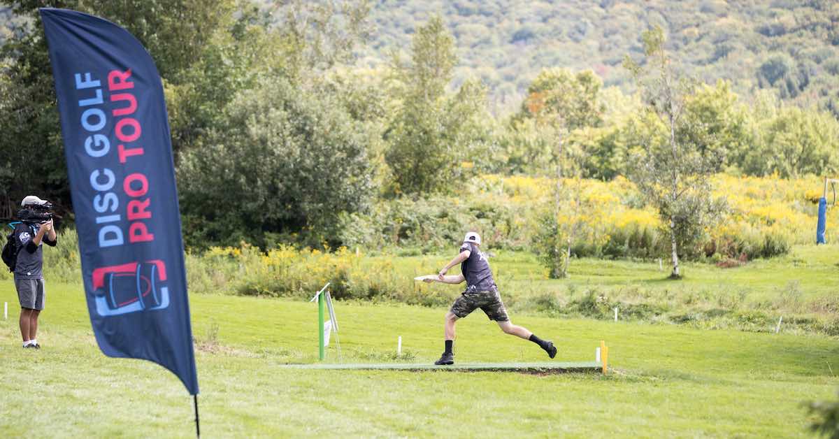 From left to right: A camerama filming, a feather banner that says "Disc Golf Pro Tour" and a disc golfer abou to drive from a tee pad