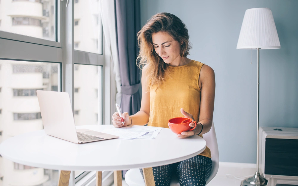 Woman writing in a notebook with a laptop in front of her
