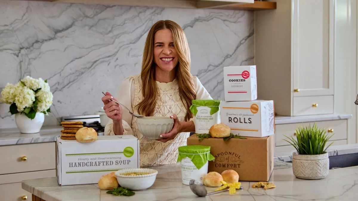 Spoonful of Comfort founder smiling in a bright kitchen while holding a bowl of soup, surrounded by handcrafted soup, rolls, cookies, and Spoonful of Comfort gift packaging.