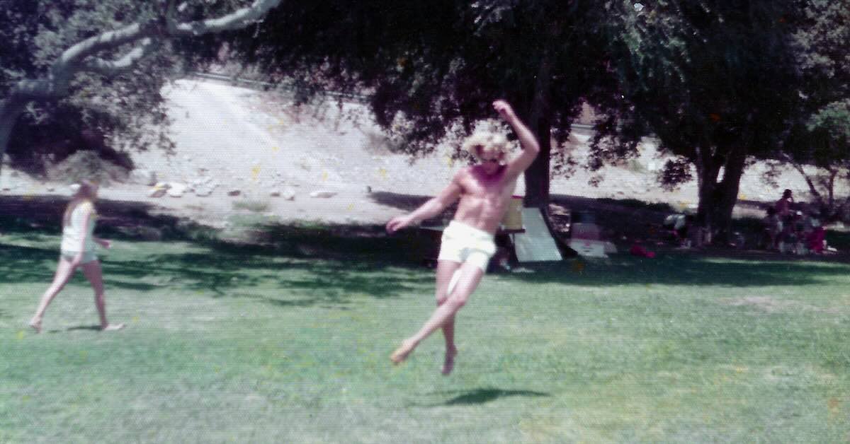 A man in just shorts catches a Frisbee with his knees in mid-air in a park