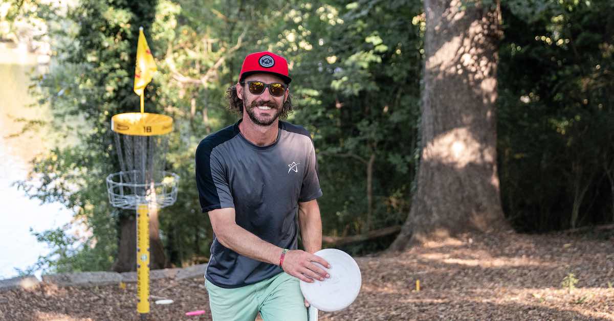 Man in hat smiling as he walks from a disc golf basket