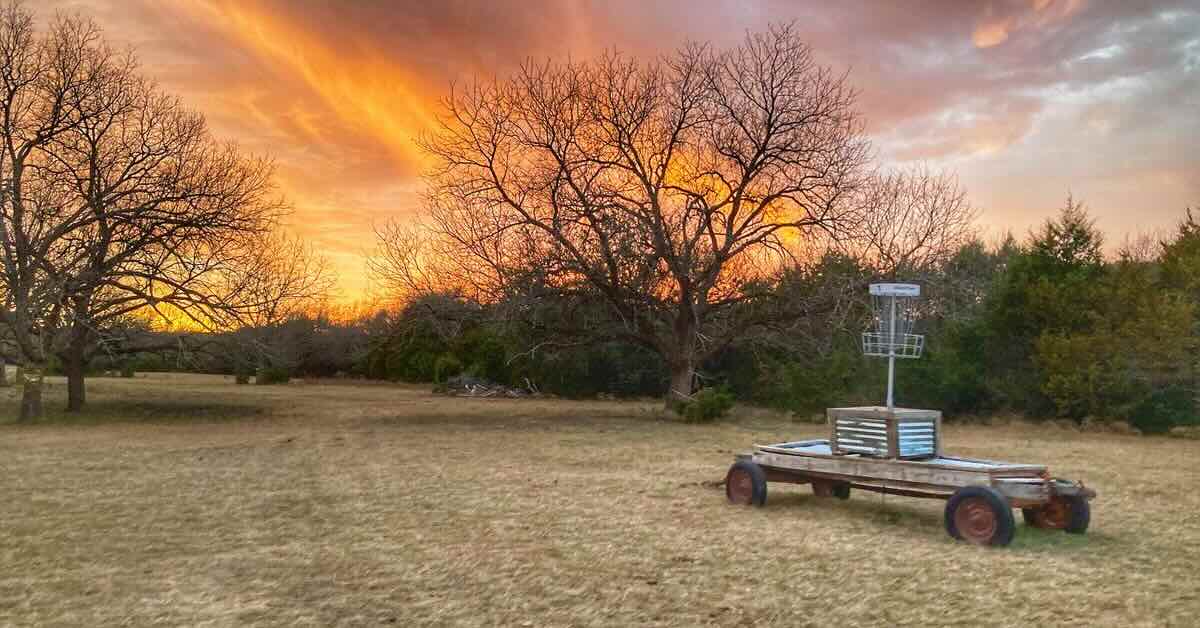 A disc golf basket mounted on an old wagon in an open area near large trees and thicker stands of small trees at sunset
