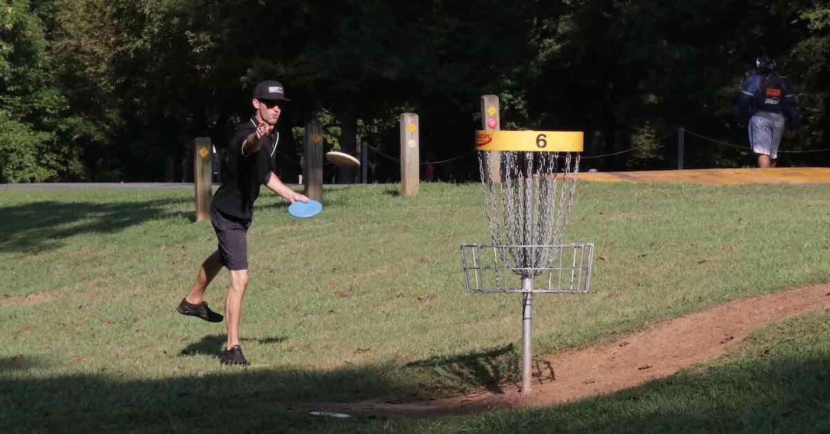 A man in a black outfit, hat, and sunglasses, putts at a disc golf basket