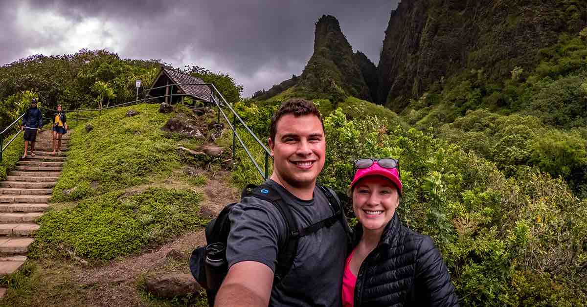 A couple smiles for a photo in front of mountains covered in lush greenery