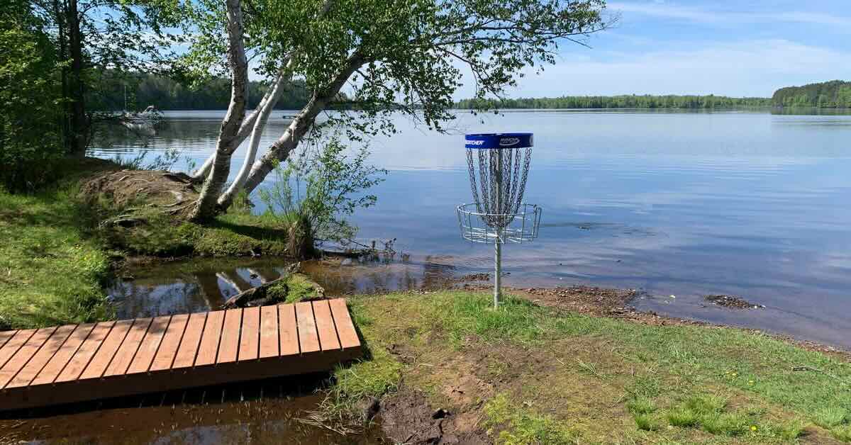 A blue banded disc golf basket on a small patch of ground next to a lake